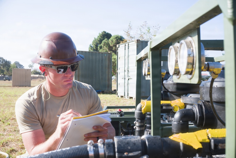 A male worker wearing a brown hard hat and sunglasses is reviewing documents on a notepad while inspecting industrial or military equipment outdoors. The equipment appears to be a green-painted pumping or fluid distribution system with multiple gauges, valves, and hoses. The setting appears to be a field or staging area with shipping containers visible in the background.