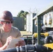 A male worker wearing a brown hard hat and sunglasses is reviewing documents on a notepad while inspecting industrial or military equipment outdoors. The equipment appears to be a green-painted pumping or fluid distribution system with multiple gauges, valves, and hoses. The setting appears to be a field or staging area with shipping containers visible in the background.