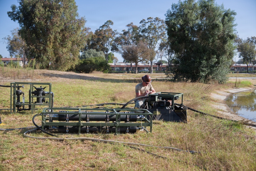 A person is working with military-green painted water pumping equipment near a small pond or water retention area. The equipment includes a pump assembly with hoses and a separate valve/manifold station, suggesting a water pumping or filtration operation. The setting appears to be a semi-arid outdoor area near institutional or military-style buildings.