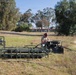 A person is working with military-green painted water pumping equipment near a small pond or water retention area. The equipment includes a pump assembly with hoses and a separate valve/manifold station, suggesting a water pumping or filtration operation. The setting appears to be a semi-arid outdoor area near institutional or military-style buildings.