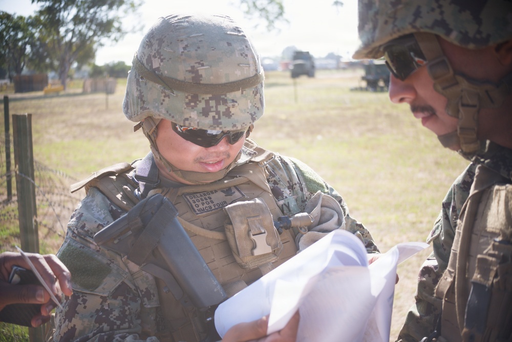Two military personnel in digital camouflage uniforms are reviewing documents or papers in an outdoor field setting. The central figure wears a tactical vest with visible name tape and unit identifiers, along with a combat helmet and sunglasses. Military vehicles and a fenced field are visible in the background.