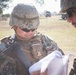 Two military personnel in digital camouflage uniforms are reviewing documents or papers in an outdoor field setting. The central figure wears a tactical vest with visible name tape and unit identifiers, along with a combat helmet and sunglasses. Military vehicles and a fenced field are visible in the background.