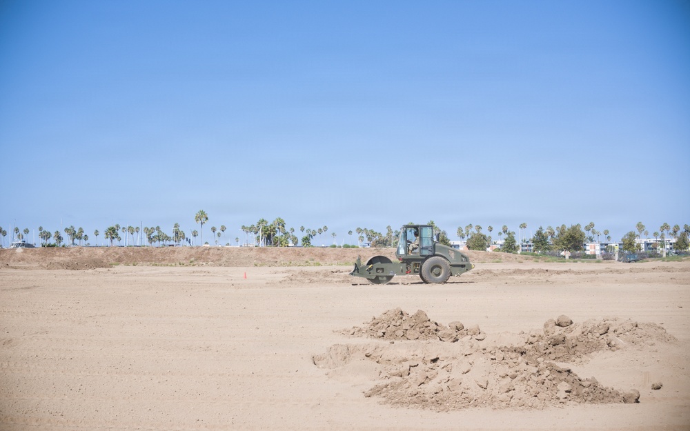 A construction site showing a soil compactor/road roller actively grading and compacting a large flat sandy area. The site appears to be in an early land preparation phase, with loose soil mounds visible in the foreground. Palm trees and low-rise buildings are visible in the background, suggesting a coastal or Southern California urban setting.