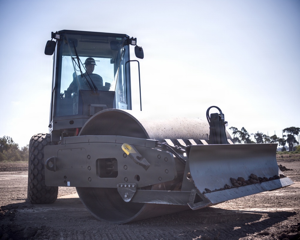 A single-drum vibratory soil compactor is being operated on a construction or grading site. The machine is equipped with a front blade/dozer attachment and a large smooth steel drum roller. An operator wearing a hard hat is visible inside the enclosed cab.