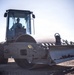 A single-drum vibratory soil compactor is being operated on a construction or grading site. The machine is equipped with a front blade/dozer attachment and a large smooth steel drum roller. An operator wearing a hard hat is visible inside the enclosed cab.