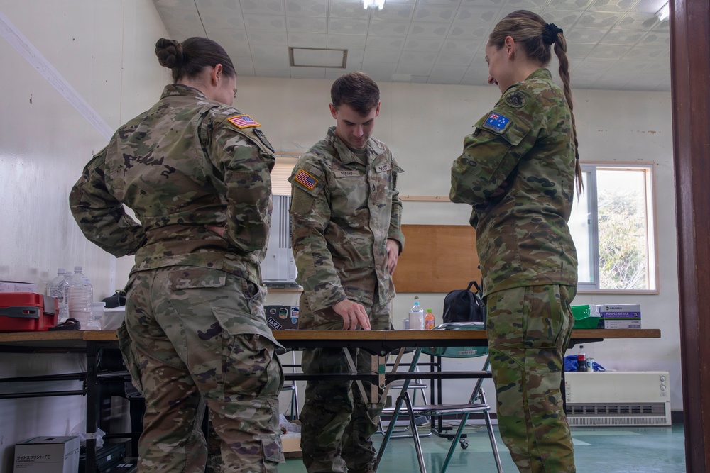 Three military personnel in camouflage uniforms are gathered around a folding table in what appears to be an indoor briefing or operations room. Two individuals wear US Army patches on their left shoulders, while the third wears an Australian flag patch, indicating a joint multinational military exercise or operation. The central figure appears to be reviewing or pointing at documents or materials on the table.