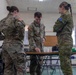 Three military personnel in camouflage uniforms are gathered around a folding table in what appears to be an indoor briefing or operations room. Two individuals wear US Army patches on their left shoulders, while the third wears an Australian flag patch, indicating a joint multinational military exercise or operation. The central figure appears to be reviewing or pointing at documents or materials on the table.