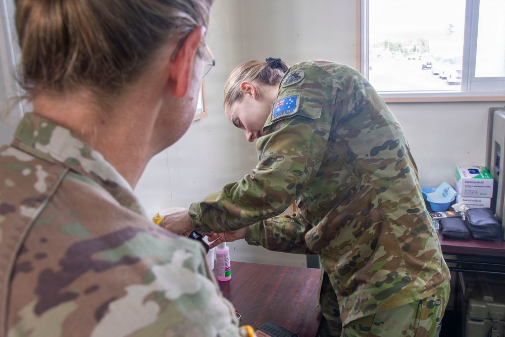 Two Australian Defence Force personnel in MultiCam camouflage uniforms are engaged in what appears to be a medical or first aid procedure at a desk or table. The soldier in the foreground has their back to the camera, while the second soldier leans forward performing a task on the first soldier's hand or wrist. The setting appears to be an indoor military medical or administrative space.