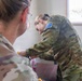 Two Australian Defence Force personnel in MultiCam camouflage uniforms are engaged in what appears to be a medical or first aid procedure at a desk or table. The soldier in the foreground has their back to the camera, while the second soldier leans forward performing a task on the first soldier's hand or wrist. The setting appears to be an indoor military medical or administrative space.