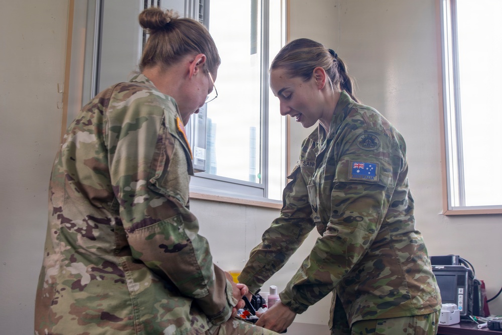 Two female military personnel in camouflage uniforms are engaged in what appears to be a medical or administrative task at a workstation inside a building. The soldier on the right wears an Australian flag patch on her right shoulder, identifying her as Australian Defence Force (ADF) personnel. They appear to be collaborating over items on a surface, possibly conducting a medical procedure or equipment check.