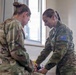 Two female military personnel in camouflage uniforms are engaged in what appears to be a medical or administrative task at a workstation inside a building. The soldier on the right wears an Australian flag patch on her right shoulder, identifying her as Australian Defence Force (ADF) personnel. They appear to be collaborating over items on a surface, possibly conducting a medical procedure or equipment check.
