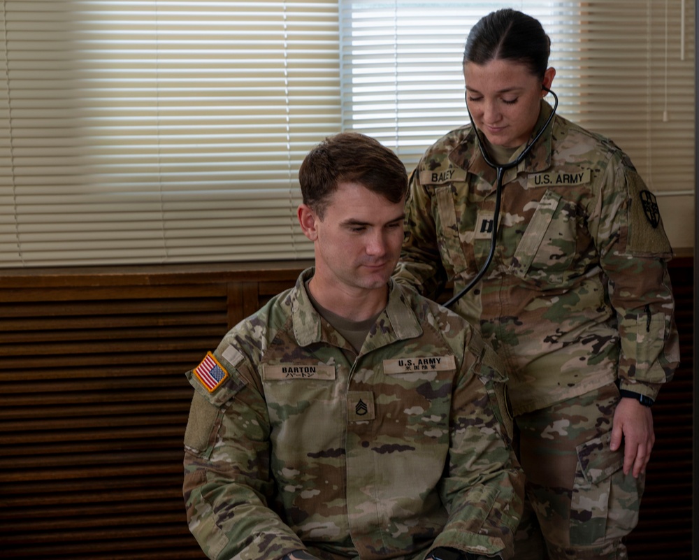 Two U.S. Army soldiers are shown in what appears to be a medical examination setting. A female soldier identified by her name tape as 'BALEY' is using a stethoscope to examine a male soldier identified as 'BARTON', who is seated. Both wear OCP (Operational Camouflage Pattern) uniforms with U.S. Army insignia.