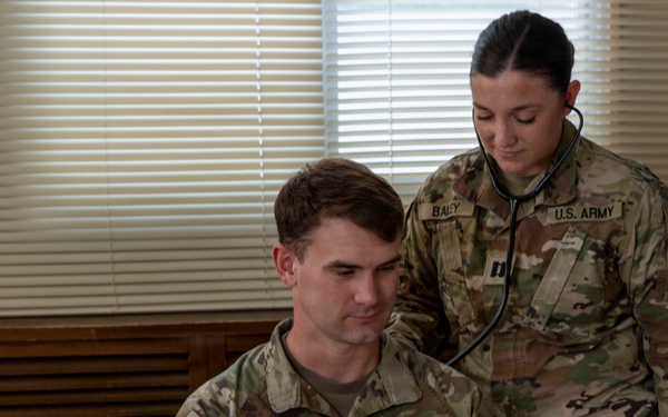 Two U.S. Army soldiers are shown in what appears to be a medical examination setting. A female soldier identified by her name tape as 'BALEY' is using a stethoscope to examine a male soldier identified as 'BARTON', who is seated. Both wear OCP (Operational Camouflage Pattern) uniforms with U.S. Army insignia.