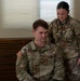 Two U.S. Army soldiers are shown in what appears to be a medical examination setting. A female soldier identified by her name tape as 'BALEY' is using a stethoscope to examine a male soldier identified as 'BARTON', who is seated. Both wear OCP (Operational Camouflage Pattern) uniforms with U.S. Army insignia.
