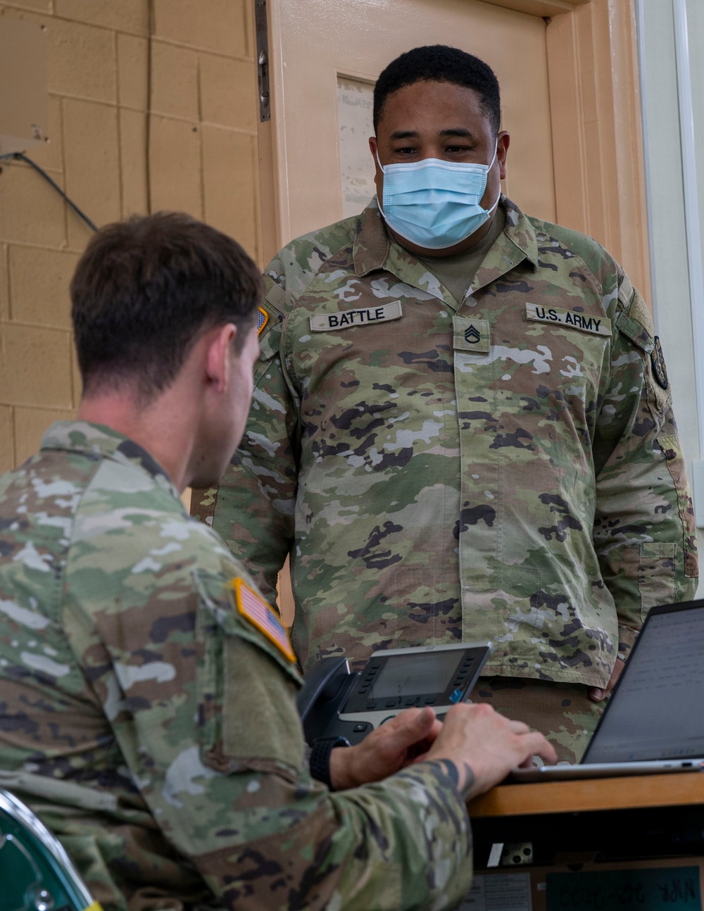Two U.S. Army soldiers in Operational Camouflage Pattern (OCP) uniforms are engaged in a work session, with one seated at a laptop and the other standing and observing. The standing soldier, identified by name tape as 'BATTLE' with U.S. ARMY branch tape, wears a blue surgical face mask and displays Staff Sergeant rank insignia. Both soldiers have U.S. flag patches on their right shoulders, consistent with Army uniform regulations.