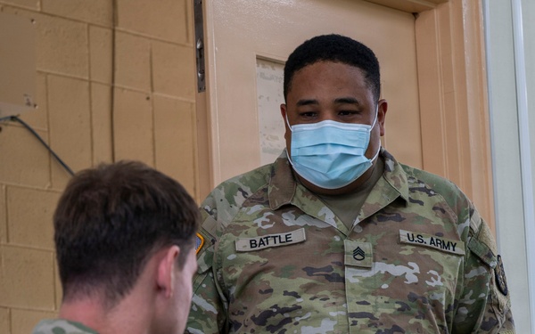Two U.S. Army soldiers in Operational Camouflage Pattern (OCP) uniforms are engaged in a work session, with one seated at a laptop and the other standing and observing. The standing soldier, identified by name tape as 'BATTLE' with U.S. ARMY branch tape, wears a blue surgical face mask and displays Staff Sergeant rank insignia. Both soldiers have U.S. flag patches on their right shoulders, consistent with Army uniform regulations.