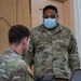 Two U.S. Army soldiers in Operational Camouflage Pattern (OCP) uniforms are engaged in a work session, with one seated at a laptop and the other standing and observing. The standing soldier, identified by name tape as 'BATTLE' with U.S. ARMY branch tape, wears a blue surgical face mask and displays Staff Sergeant rank insignia. Both soldiers have U.S. flag patches on their right shoulders, consistent with Army uniform regulations.