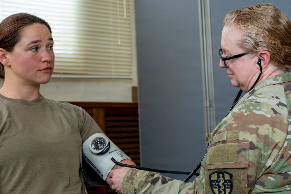 A military medical personnel in MultiCam/OCP camouflage uniform is taking the blood pressure of another service member wearing an olive drab t-shirt. The medical provider is using a Welch Allyn sphygmomanometer (blood pressure cuff) and a stethoscope. The setting appears to be a military medical facility or examination room.