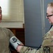 A military medical personnel in MultiCam/OCP camouflage uniform is taking the blood pressure of another service member wearing an olive drab t-shirt. The medical provider is using a Welch Allyn sphygmomanometer (blood pressure cuff) and a stethoscope. The setting appears to be a military medical facility or examination room.