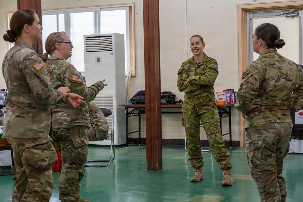 Four female military personnel in camouflage uniforms are engaged in a casual conversation inside what appears to be a military facility or base building. Two of the individuals wear US Army patches (American flag on sleeve), while the central figure wears a distinctly different camouflage pattern consistent with Australian Defence Force (ADF) uniform with a visible Australian flag patch. The interaction appears relaxed and informal, suggesting a multinational military cooperation or joint exercise setting.
