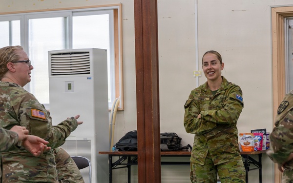 Four female military personnel in camouflage uniforms are engaged in a casual conversation inside what appears to be a military facility or base building. Two of the individuals wear US Army patches (American flag on sleeve), while the central figure wears a distinctly different camouflage pattern consistent with Australian Defence Force (ADF) uniform with a visible Australian flag patch. The interaction appears relaxed and informal, suggesting a multinational military cooperation or joint exercise setting.