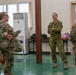 Four female military personnel in camouflage uniforms are engaged in a casual conversation inside what appears to be a military facility or base building. Two of the individuals wear US Army patches (American flag on sleeve), while the central figure wears a distinctly different camouflage pattern consistent with Australian Defence Force (ADF) uniform with a visible Australian flag patch. The interaction appears relaxed and informal, suggesting a multinational military cooperation or joint exercise setting.