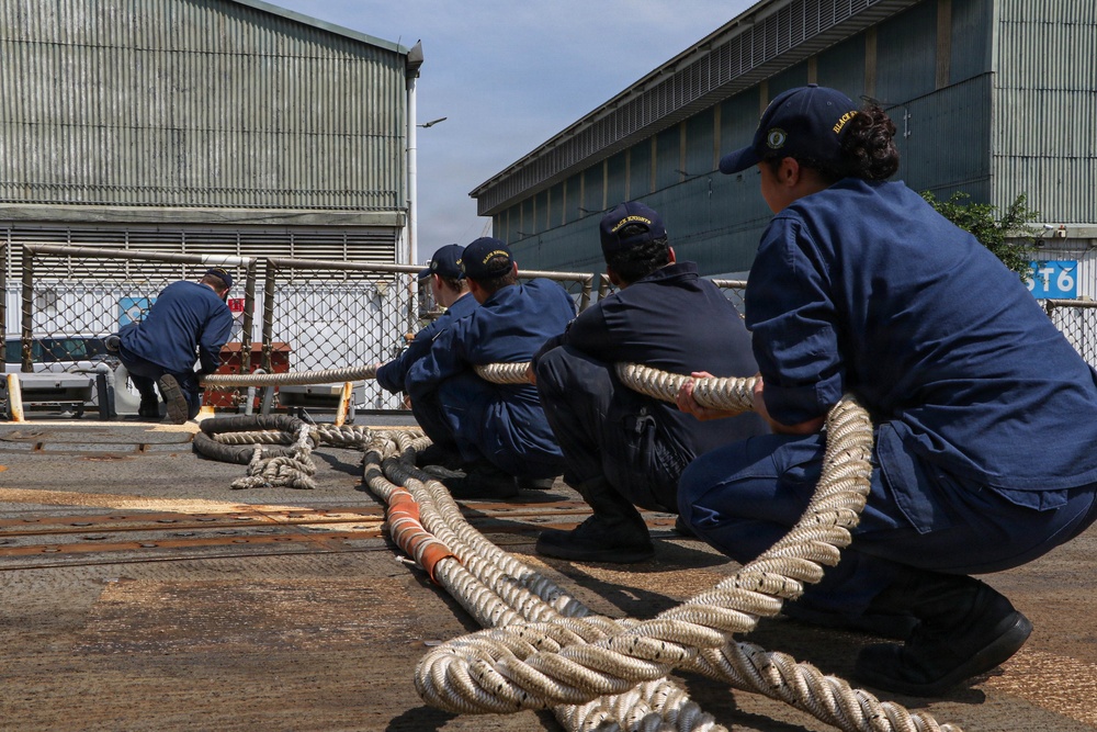 A group of personnel dressed in matching navy blue uniforms and caps are crouching in a line, collectively pulling on a large thick rope in what appears to be a port or industrial dockyard setting. The coordinated effort suggests a mooring or line-handling exercise, likely aboard or near a vessel. Large corrugated metal warehouse buildings are visible in the background.