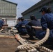 A group of personnel dressed in matching navy blue uniforms and caps are crouching in a line, collectively pulling on a large thick rope in what appears to be a port or industrial dockyard setting. The coordinated effort suggests a mooring or line-handling exercise, likely aboard or near a vessel. Large corrugated metal warehouse buildings are visible in the background.