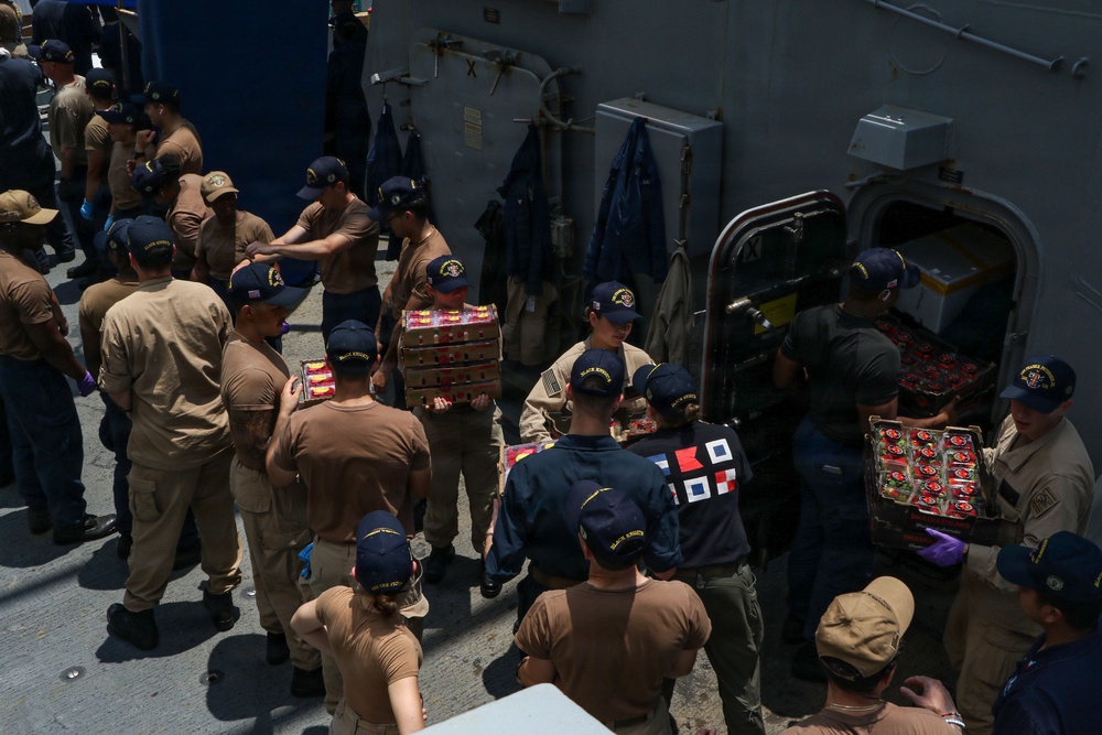 A group of military personnel, appearing to be U.S. Navy sailors, are conducting a stores onload or replenishment operation on the deck of a naval vessel. They are forming a human chain to pass trays of what appear to be fresh produce (likely strawberries or tomatoes) through a hatch into the ship's interior. The personnel are wearing standard Navy working uniforms (NWUs) and unit-specific ball caps.