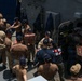 A group of military personnel, appearing to be U.S. Navy sailors, are conducting a stores onload or replenishment operation on the deck of a naval vessel. They are forming a human chain to pass trays of what appear to be fresh produce (likely strawberries or tomatoes) through a hatch into the ship's interior. The personnel are wearing standard Navy working uniforms (NWUs) and unit-specific ball caps.