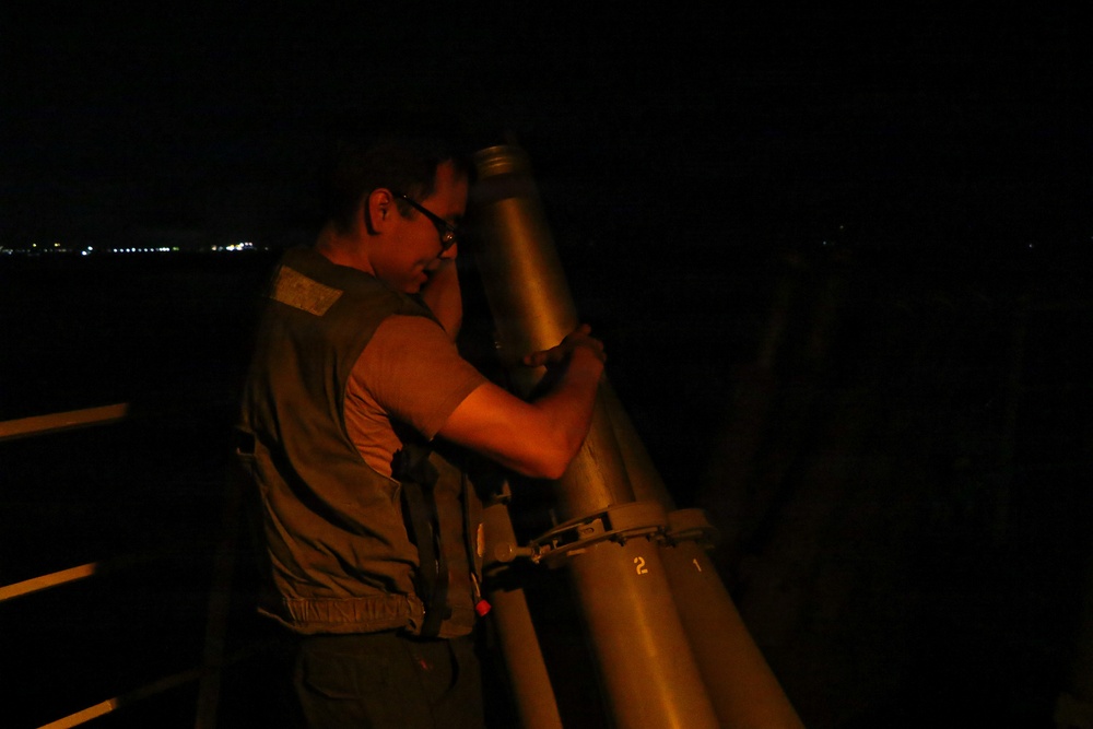 A man wearing glasses and a vest is looking through a large mounted telescope or observation device at night. The scene appears to be on an elevated platform or observation deck, with distant city lights visible on the horizon to the left. The image is taken in low-light conditions with warm artificial lighting illuminating the subject.