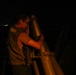 A man wearing glasses and a vest is looking through a large mounted telescope or observation device at night. The scene appears to be on an elevated platform or observation deck, with distant city lights visible on the horizon to the left. The image is taken in low-light conditions with warm artificial lighting illuminating the subject.