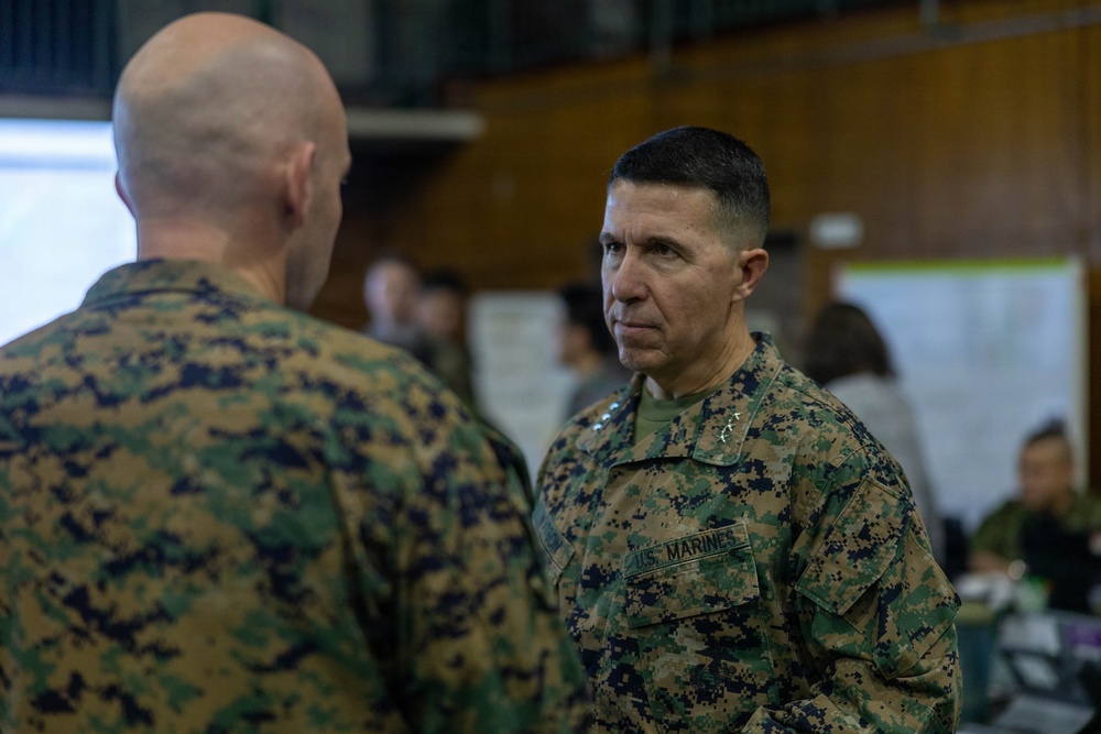 Two U.S. Marines in MARPAT digital camouflage uniforms are engaged in a face-to-face conversation in what appears to be a military operations or briefing room. The Marine facing the camera displays rank insignia consistent with a general officer (two stars visible on collar) and a 'U.S. MARINES' chest tape. The background shows additional personnel working at tables with documents and charts posted on walls.