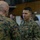 Two U.S. Marines in MARPAT digital camouflage uniforms are engaged in a face-to-face conversation in what appears to be a military operations or briefing room. The Marine facing the camera displays rank insignia consistent with a general officer (two stars visible on collar) and a 'U.S. MARINES' chest tape. The background shows additional personnel working at tables with documents and charts posted on walls.