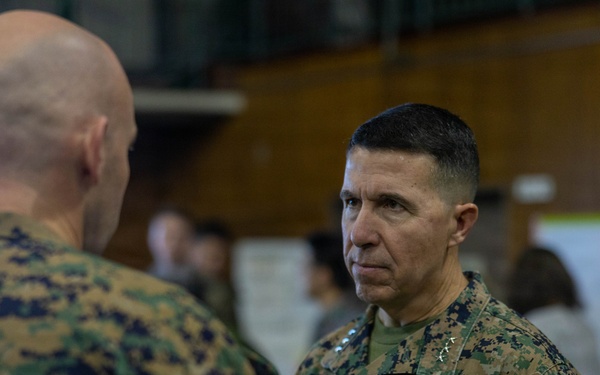 Two U.S. Marines in MARPAT digital camouflage uniforms are engaged in a face-to-face conversation in what appears to be a military operations or briefing room. The Marine facing the camera displays rank insignia consistent with a general officer (two stars visible on collar) and a 'U.S. MARINES' chest tape. The background shows additional personnel working at tables with documents and charts posted on walls.