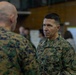 Two U.S. Marines in MARPAT digital camouflage uniforms are engaged in a face-to-face conversation in what appears to be a military operations or briefing room. The Marine facing the camera displays rank insignia consistent with a general officer (two stars visible on collar) and a 'U.S. MARINES' chest tape. The background shows additional personnel working at tables with documents and charts posted on walls.