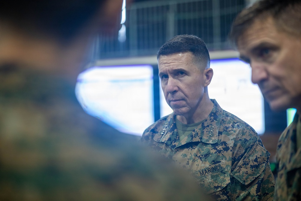 A U.S. Marine Corps officer in the foreground wearing MARPAT digital camouflage uniform with 'MARINES' tape and what appears to be general officer rank insignia (stars) on the collar. He is in what appears to be a command operations center or briefing room with illuminated screens visible in the background. At least two other military personnel are partially visible flanking him.