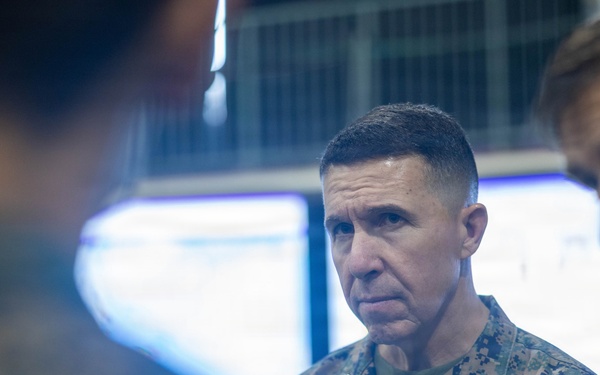 A U.S. Marine Corps officer in the foreground wearing MARPAT digital camouflage uniform with 'MARINES' tape and what appears to be general officer rank insignia (stars) on the collar. He is in what appears to be a command operations center or briefing room with illuminated screens visible in the background. At least two other military personnel are partially visible flanking him.