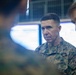 A U.S. Marine Corps officer in the foreground wearing MARPAT digital camouflage uniform with 'MARINES' tape and what appears to be general officer rank insignia (stars) on the collar. He is in what appears to be a command operations center or briefing room with illuminated screens visible in the background. At least two other military personnel are partially visible flanking him.