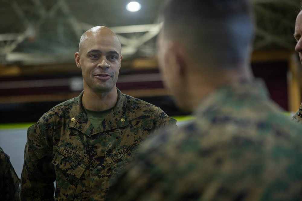 A U.S. Marine officer with the name tape 'MARTINEZ' and gold oak leaf cluster insignia (indicating the rank of Major) is engaged in conversation with other Marines in what appears to be an indoor military facility or gymnasium. He is wearing the standard Marine Corps MARPAT woodland camouflage uniform. The image captures an informal briefing or discussion among military personnel.