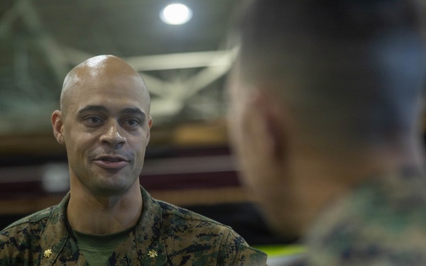 A U.S. Marine officer with the name tape 'MARTINEZ' and gold oak leaf cluster insignia (indicating the rank of Major) is engaged in conversation with other Marines in what appears to be an indoor military facility or gymnasium. He is wearing the standard Marine Corps MARPAT woodland camouflage uniform. The image captures an informal briefing or discussion among military personnel.