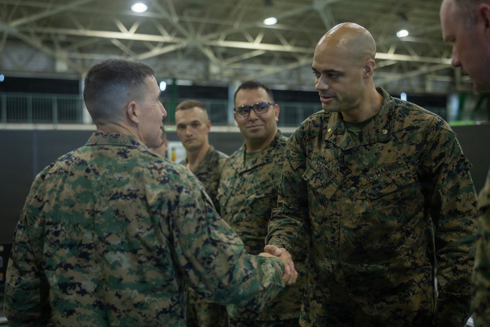Several U.S. Marines in MARPAT woodland camouflage uniforms are gathered in what appears to be a large indoor facility, possibly a hangar or gymnasium. Two Marines in the foreground are engaged in a handshake, suggesting a formal greeting, recognition, or introduction ceremony. Additional Marines observe the interaction in the background.