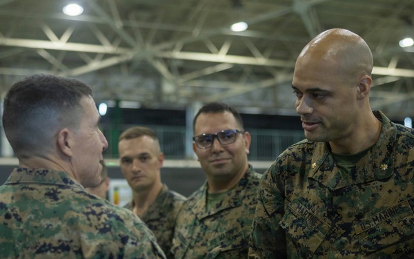 Several U.S. Marines in MARPAT woodland camouflage uniforms are gathered in what appears to be a large indoor facility, possibly a hangar or gymnasium. Two Marines in the foreground are engaged in a handshake, suggesting a formal greeting, recognition, or introduction ceremony. Additional Marines observe the interaction in the background.