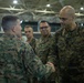 Several U.S. Marines in MARPAT woodland camouflage uniforms are gathered in what appears to be a large indoor facility, possibly a hangar or gymnasium. Two Marines in the foreground are engaged in a handshake, suggesting a formal greeting, recognition, or introduction ceremony. Additional Marines observe the interaction in the background.