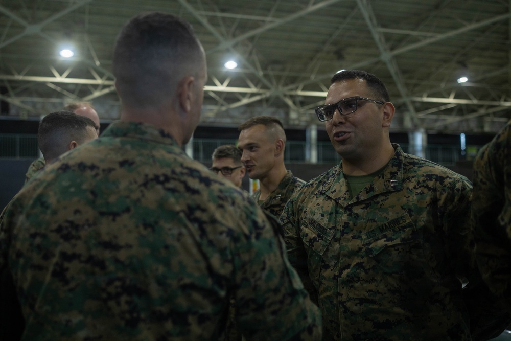 A group of U.S. Marines in MARPAT digital camouflage uniforms are gathered inside what appears to be a large hangar or indoor facility. The central focus is on two Marines engaged in conversation, with one facing the camera wearing glasses and a name tape reading 'US MARINES'. Several other Marines are visible in the background.