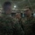 A group of U.S. Marines in MARPAT digital camouflage uniforms are gathered inside what appears to be a large hangar or indoor facility. The central focus is on two Marines engaged in conversation, with one facing the camera wearing glasses and a name tape reading 'US MARINES'. Several other Marines are visible in the background.