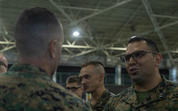 A group of U.S. Marines in MARPAT digital camouflage uniforms are gathered inside what appears to be a large hangar or indoor facility. The central focus is on two Marines engaged in conversation, with one facing the camera wearing glasses and a name tape reading 'US MARINES'. Several other Marines are visible in the background.