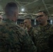 A group of U.S. Marines in MARPAT digital camouflage uniforms are gathered inside what appears to be a large hangar or indoor facility. The central focus is on two Marines engaged in conversation, with one facing the camera wearing glasses and a name tape reading 'US MARINES'. Several other Marines are visible in the background.