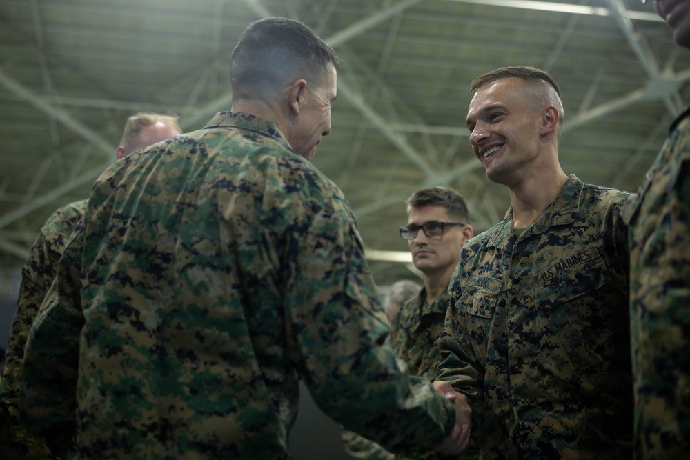 Two U.S. Marines in MARPAT camouflage uniforms are shaking hands in what appears to be a hangar or large indoor military facility. The Marine on the right is smiling broadly, suggesting a positive interaction such as a commendation, award, or greeting ceremony. Several other Marines are visible in the background observing the exchange.