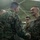 Two U.S. Marines in MARPAT camouflage uniforms are shaking hands in what appears to be a hangar or large indoor military facility. The Marine on the right is smiling broadly, suggesting a positive interaction such as a commendation, award, or greeting ceremony. Several other Marines are visible in the background observing the exchange.